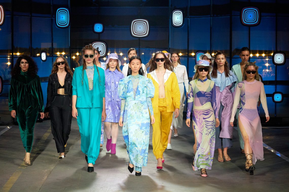 Un groupe de mannequins défilant sur un podium lors d'un défilé de mode, portant des tenues colorées et variées. Les vêtements incluent des costumes, des robes et des ensembles assortis dans des tons vifs comme le turquoise, le jaune, le violet, et le vert. L'arrière-plan est décoré avec des motifs géométriques lumineux.