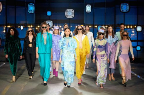 Un groupe de mannequins défilant sur un podium lors d'un défilé de mode, portant des tenues colorées et variées. Les vêtements incluent des costumes, des robes et des ensembles assortis dans des tons vifs comme le turquoise, le jaune, le violet, et le vert. L'arrière-plan est décoré avec des motifs géométriques lumineux.