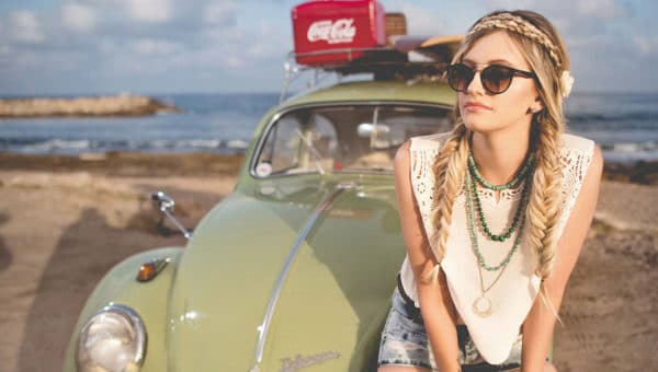 Une femme avec des lunettes de soleil et des cheveux blonds tressés, portant un haut blanc et un short en jean, est assise sur le capot d'une voiture verte avec une planche de surf sur le toit. La voiture est garée près de la plage avec la mer en arrière-plan.
