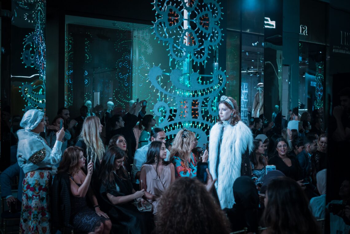 Un défilé de mode se déroulant dans un espace décoré avec des lumières bleues et des éléments futuristes. Un mannequin portant une veste blanche à fourrure et un diadème marche sur le podium, entouré d'un public prenant des photos et des vidéos. L'ambiance est festive et animée.