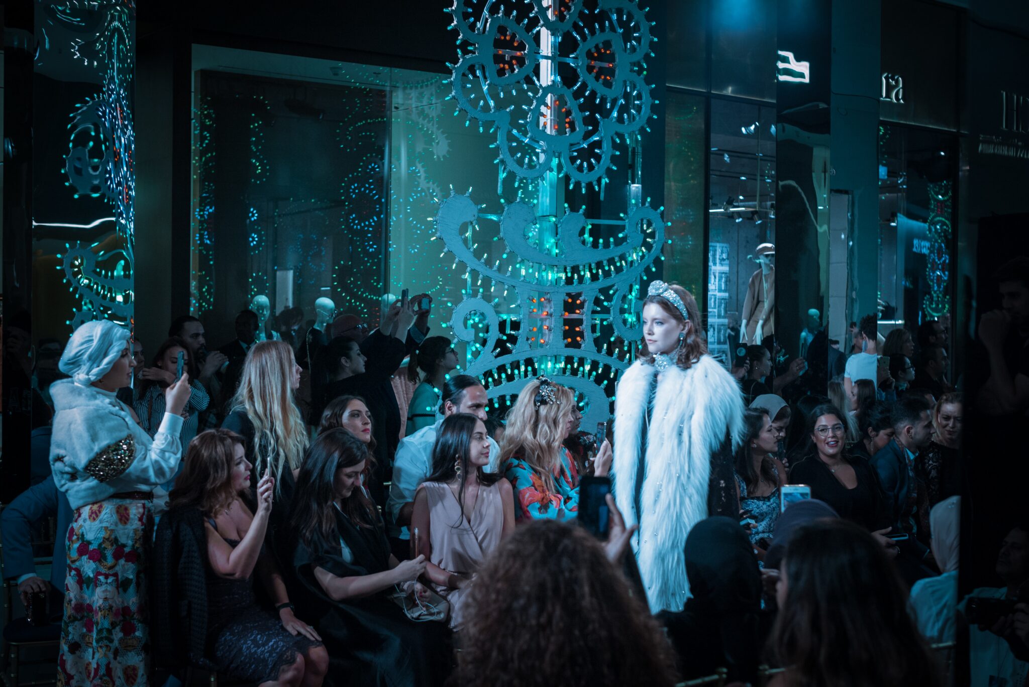 Un défilé de mode se déroulant dans un espace décoré avec des lumières bleues et des éléments futuristes. Un mannequin portant une veste blanche à fourrure et un diadème marche sur le podium, entouré d'un public prenant des photos et des vidéos. L'ambiance est festive et animée.