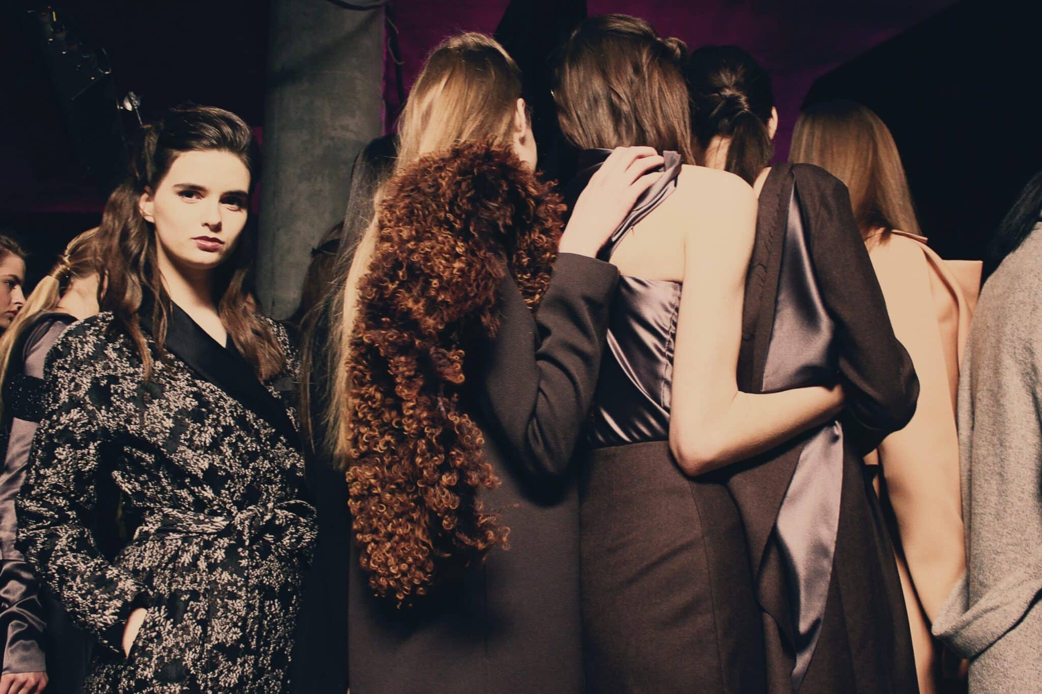 Groupe de mannequins en coulisses lors d'un défilé de mode, portant des tenues élégantes et variées, avec des textures et des styles distincts, incluant des robes et des vestes.