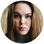 Portrait d'une femme avec des cheveux blonds et un regard sérieux. Elle a les sourcils légèrement froncés et les lèvres légèrement entrouvertes. Le fond est flou, mettant l'accent sur son visage.